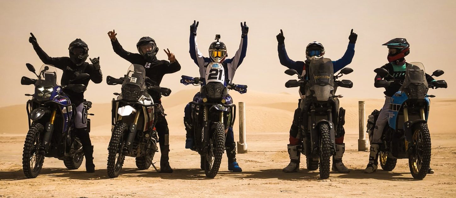 Cinque motociclisti in posa con le braccia alzate davanti alle dune del Sahara