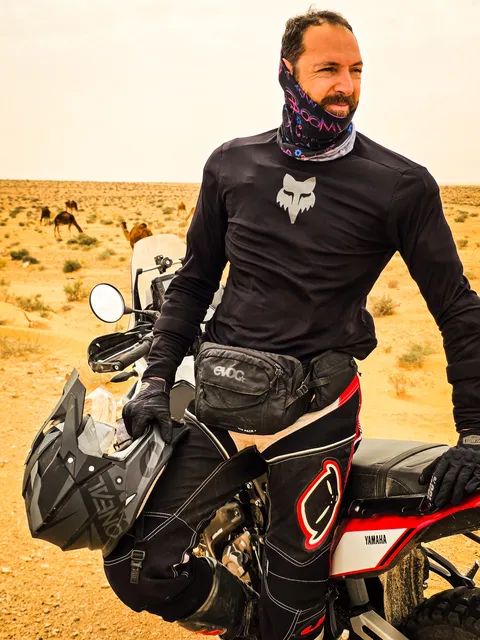 Ritratto di un pilota appoggiato alla moto, con i cammelli nel deserto alle spalle