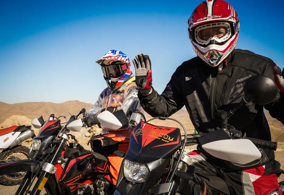 Due piloti salutano dalle moto, con le colline del deserto dietro